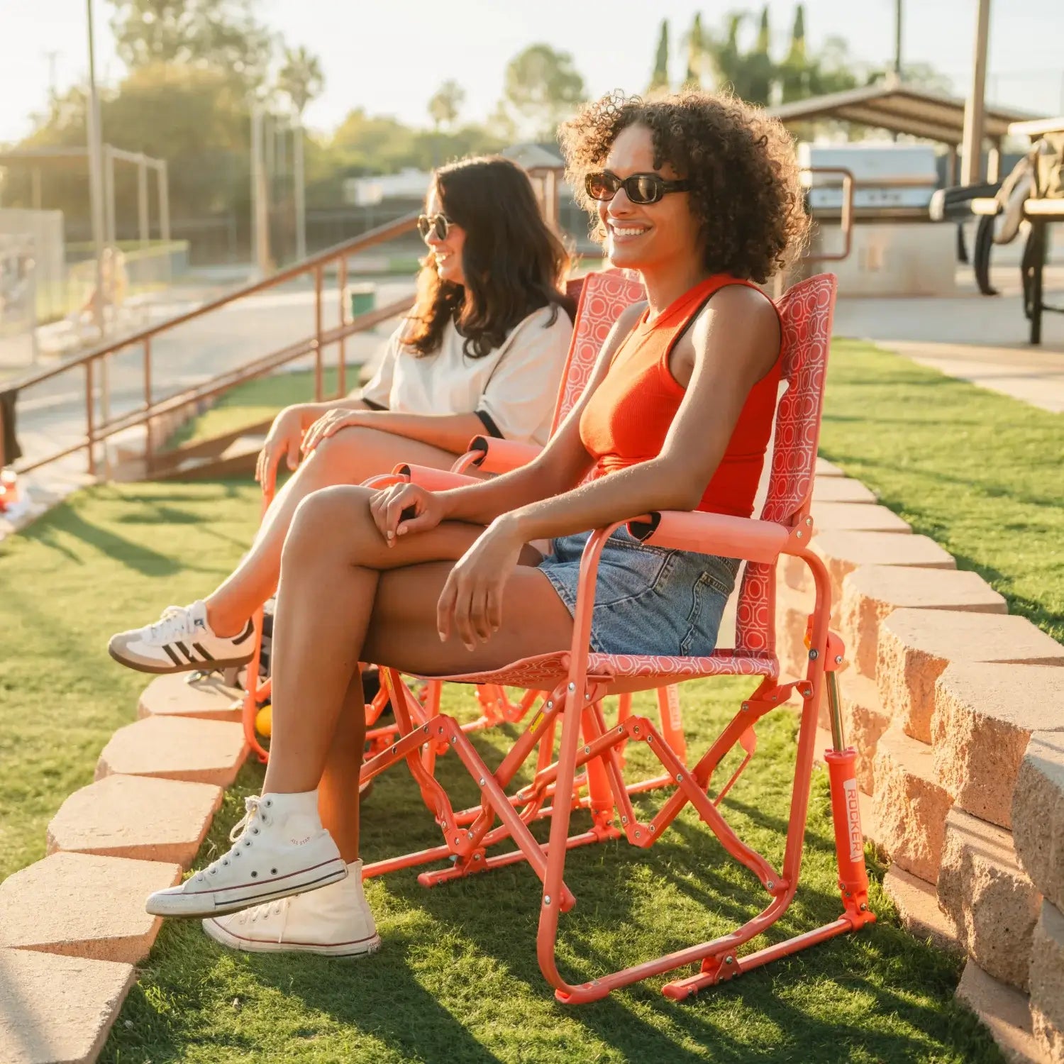 Two women sitting outdoors in GCI Outdoor Stowaway Rockers in Coral Mosaic, enjoying a sunny day at a sports field.