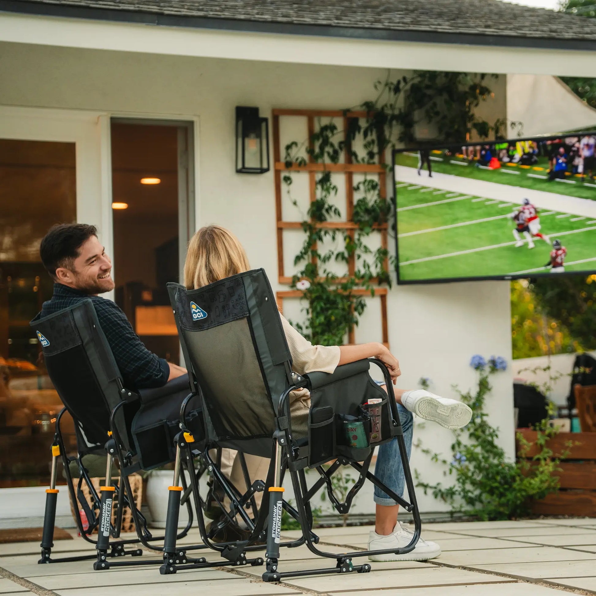 Two people sitting in tailgater dark charcoal Stowaway Rocker chairs while watching a football game in a backyard. 