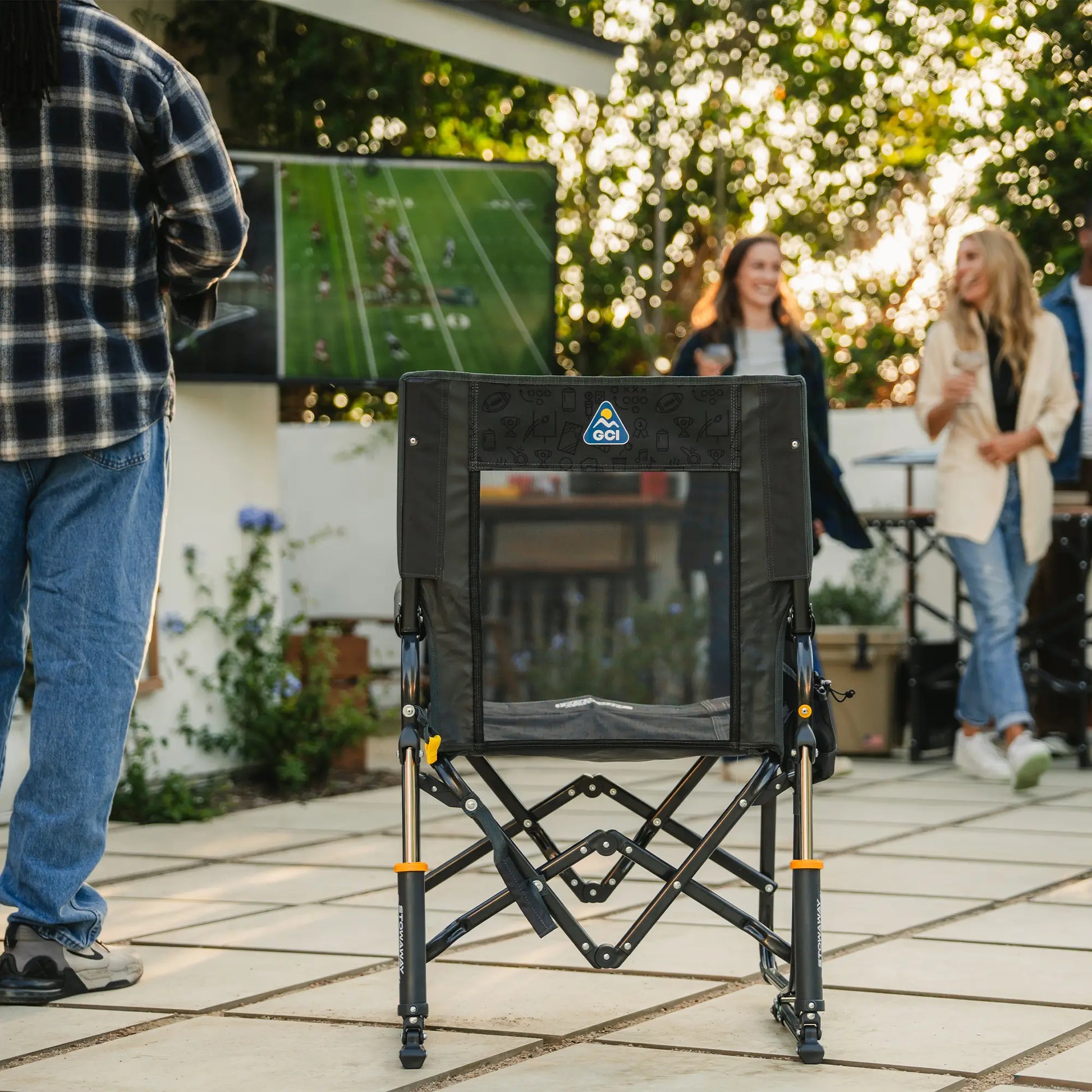 A tailgater dark charcoal Stowaway Rocker positioned in a backyard setting with people standing around watching television. 