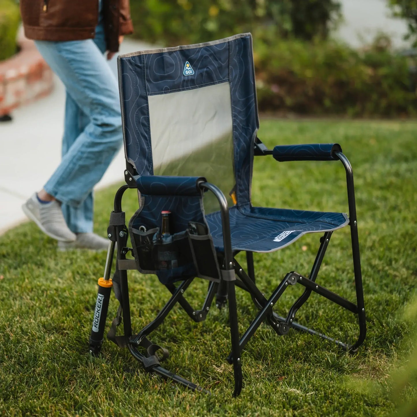 A navy topo Stowaway Rocker unfolded on a patch of grass while a person walks closely behind the chair. 