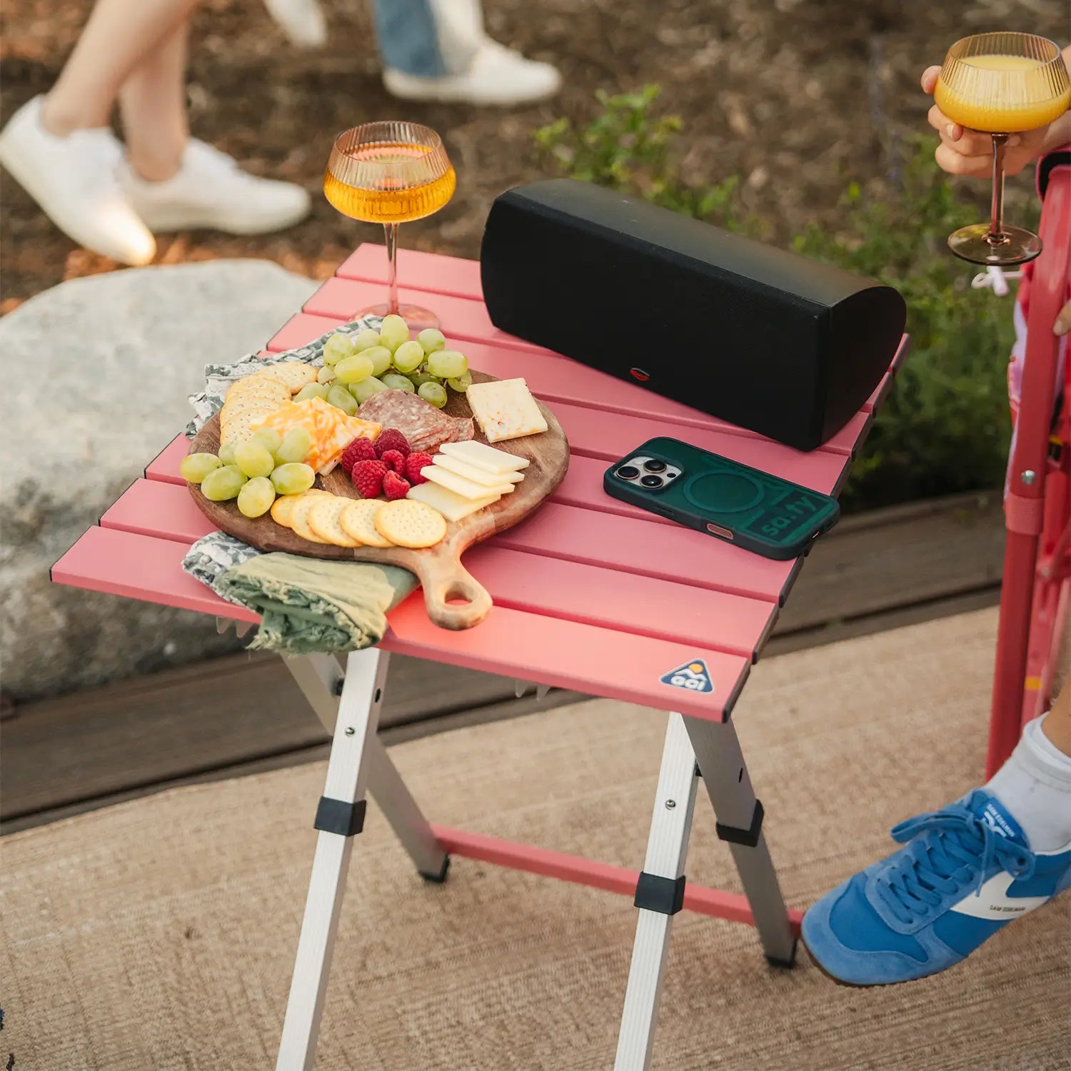 A speaker and charcuterie tray resting on a coral Compact Camp Table. 