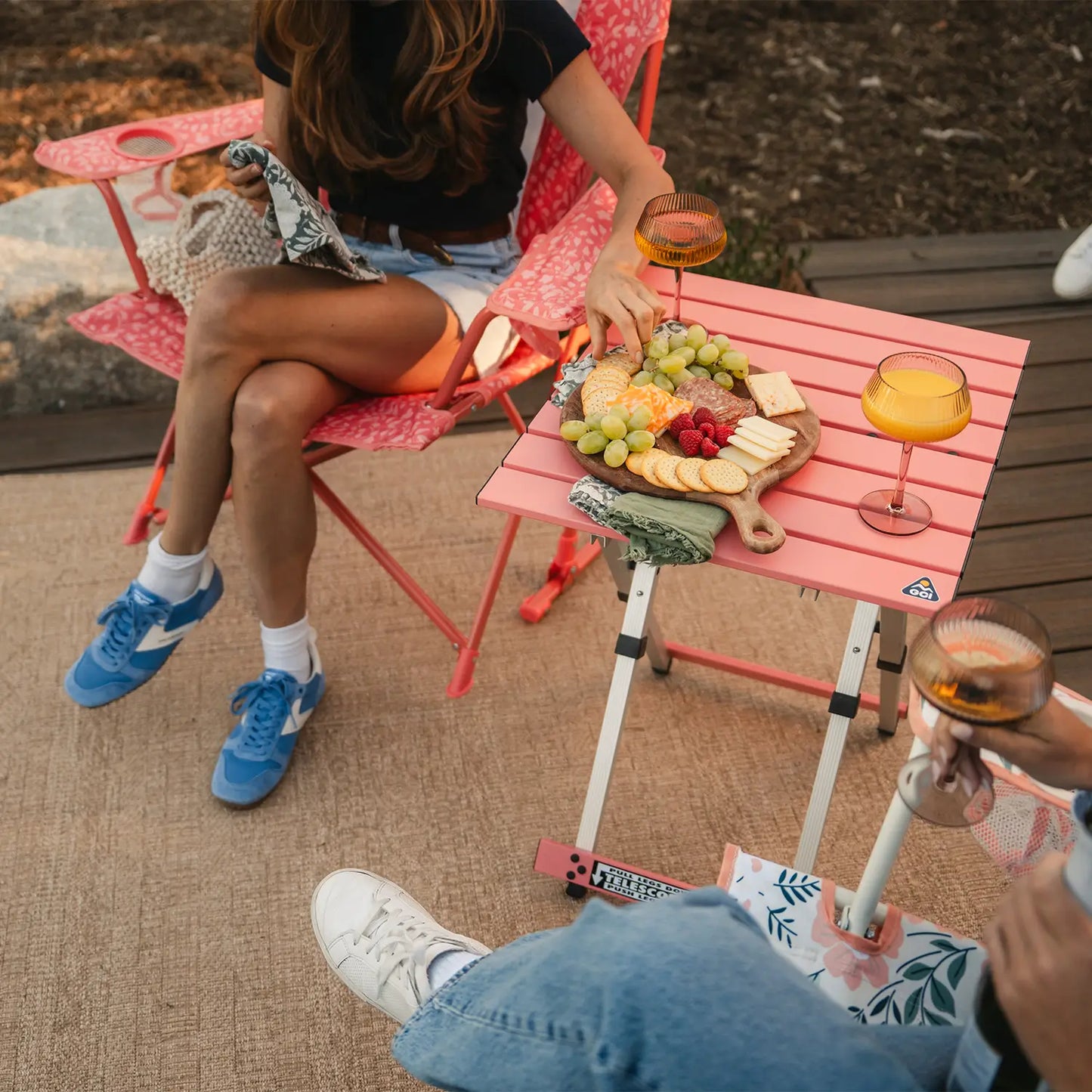 Two women sitting in coral Comfort Pro Rockers and having a snack thats positioned on a coral Compact Camp Table. 