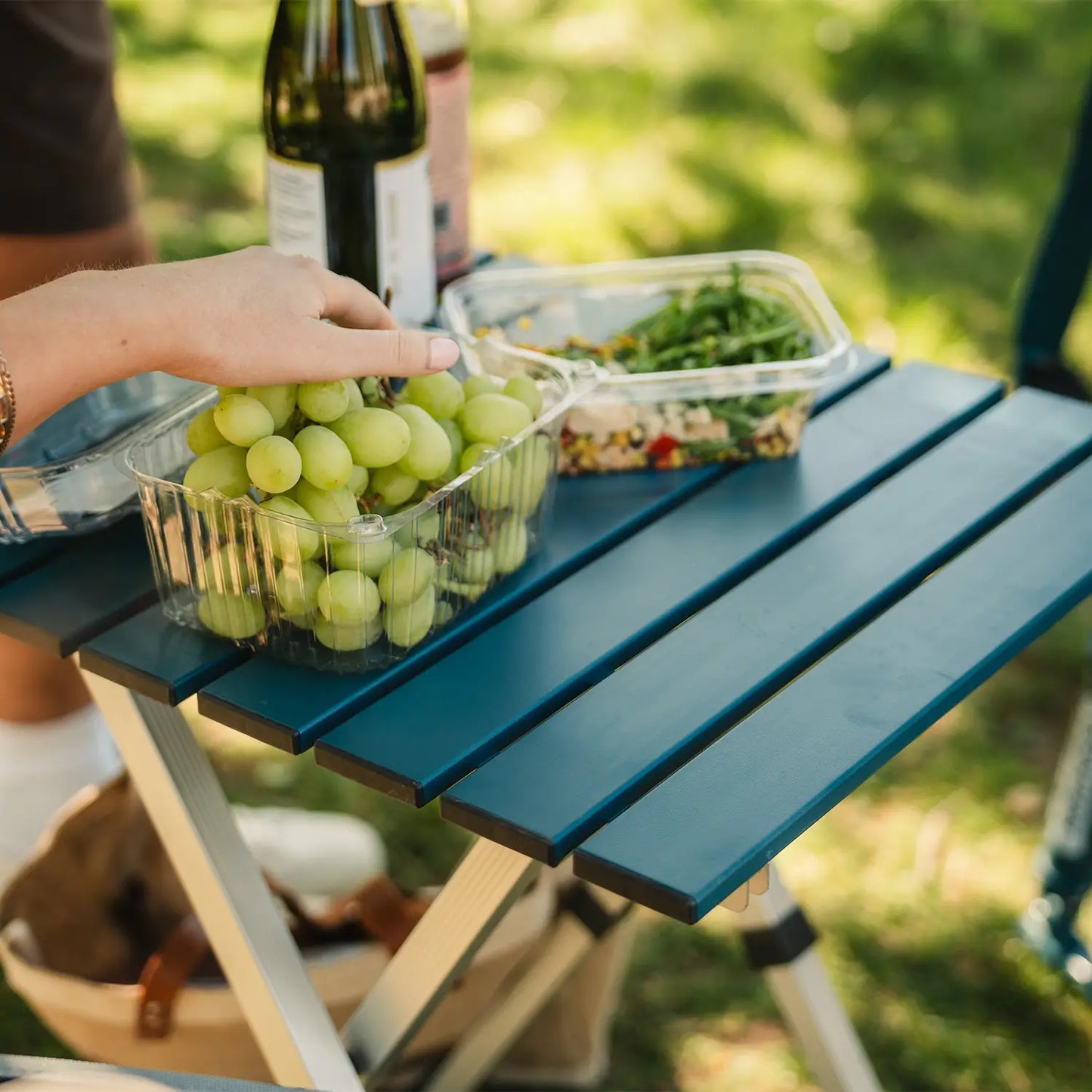 A person reaching for a pack of grapes that are positioned on a teal Compact Camp Table. 