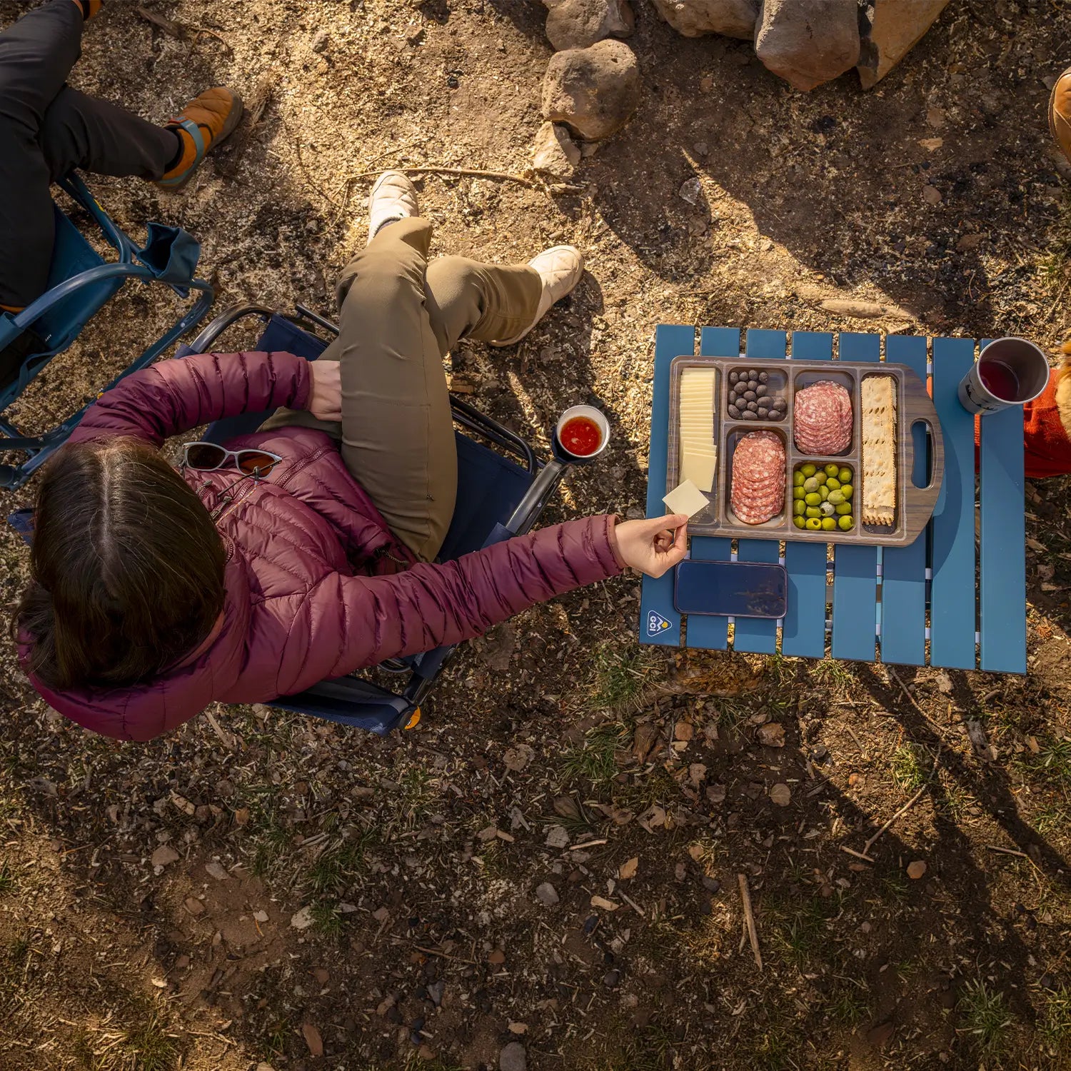 An aerial view of a woman eating cheese from a charcuterie on top of a teal Compact Camp Table. 