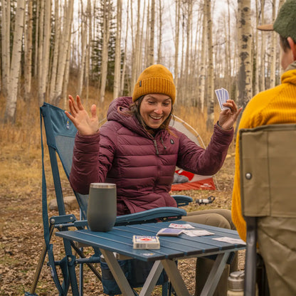 A couple playing cards on a teal Compact Camp Table while they camp in the woods. 