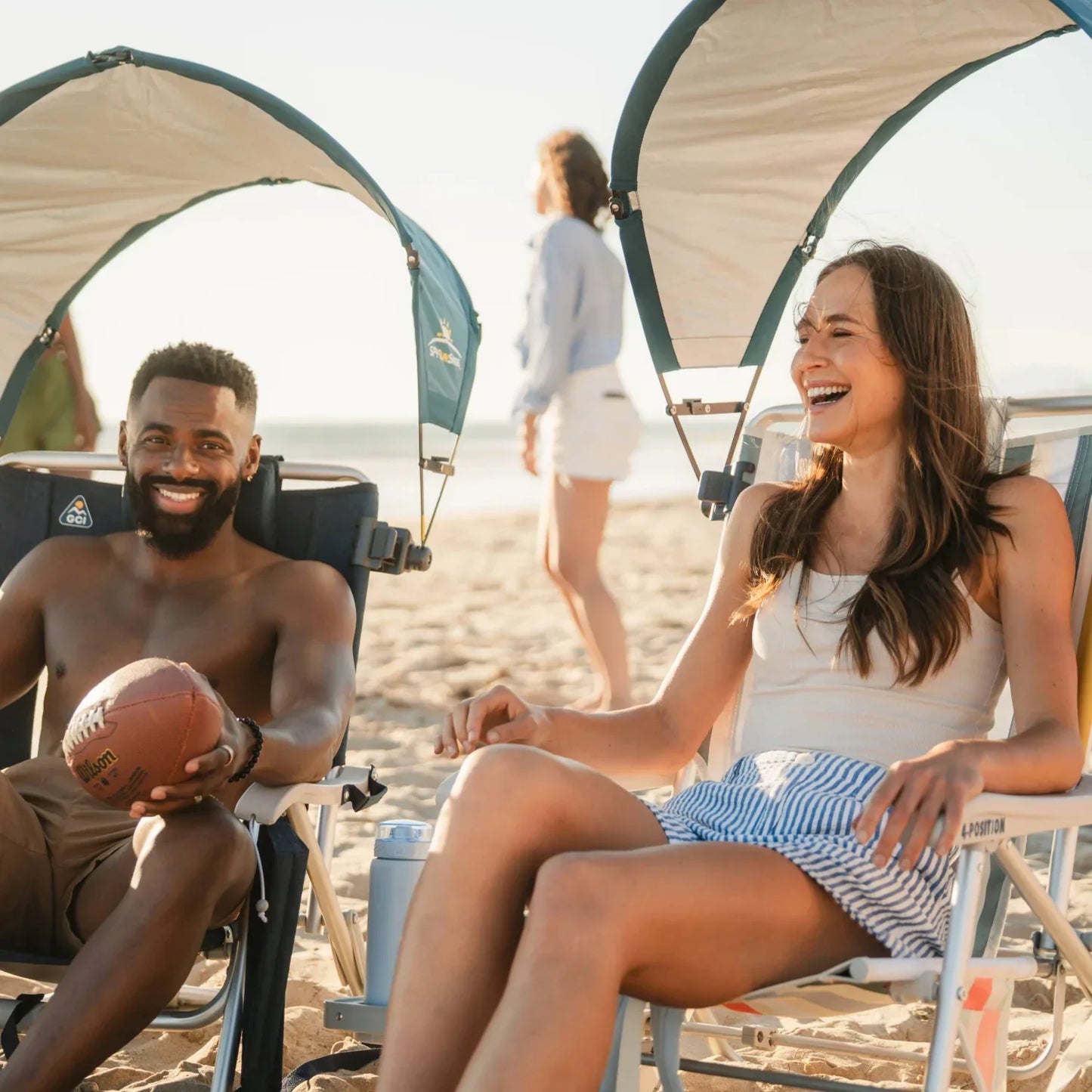 A woman and a man laughing in beach chairs fitted with teal GCI Outdoor SPF SunShade Accessories, the man holding a football.