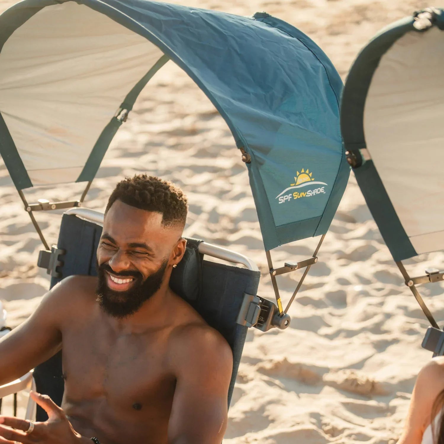 Man smiling beneath the arched canopy of a teal GCI Outdoor SPF SunShade Accessory clamped to his beach chair.