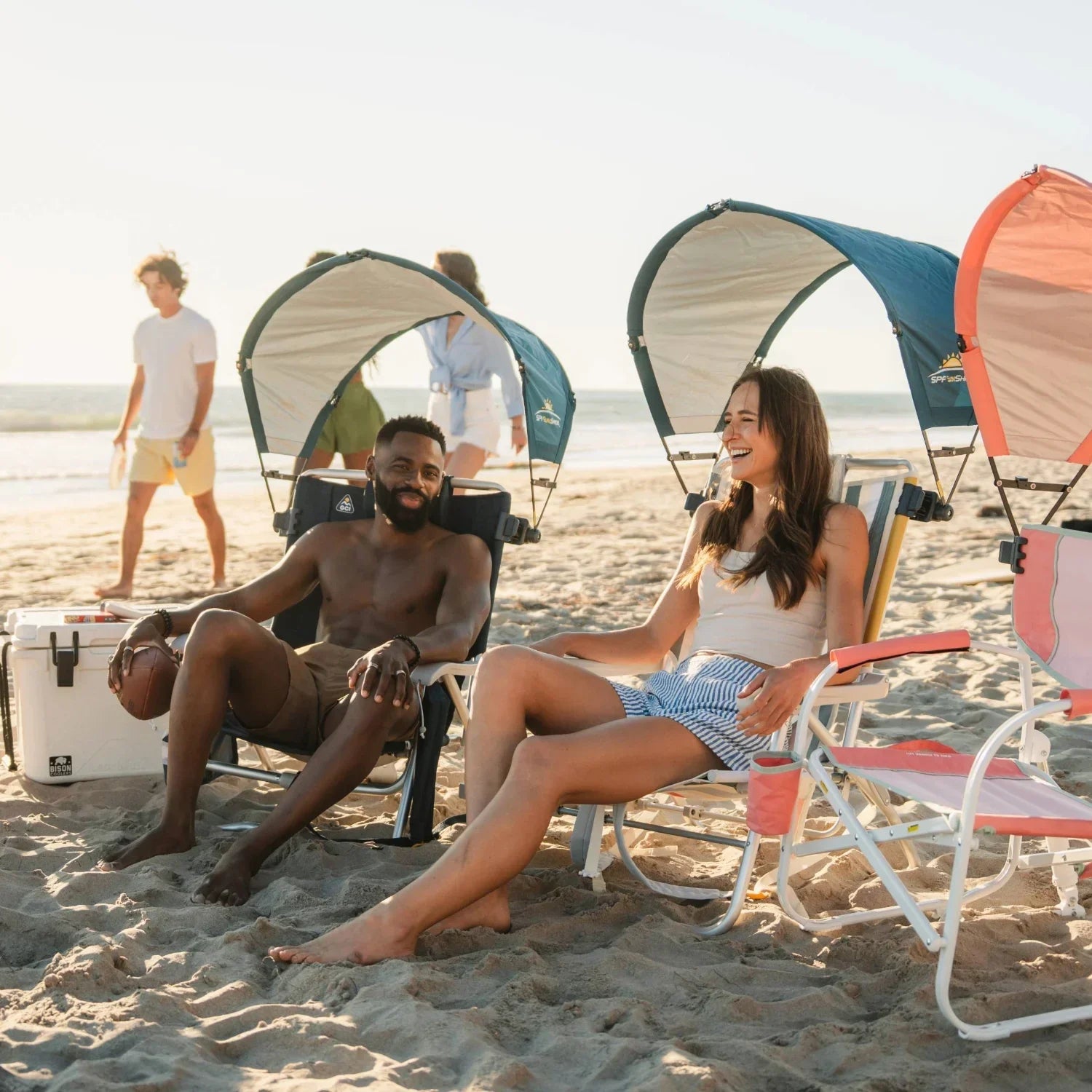 Friends on the beach using chairs with teal and coral GCI Outdoor SPF SunShade Accessories, showing color options in use.