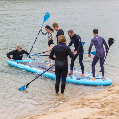 Group of six people on a large blue inflatable paddleboard in shallow water, some paddling, others balancing.