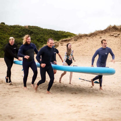 Group of six people in wetsuits running on sand with a large blue paddleboard