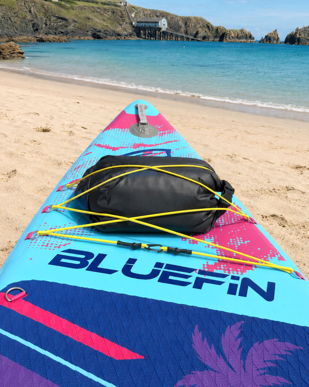 Inflatable paddleboard on beach with black dry bag secured by yellow bungee cords, blue water in background