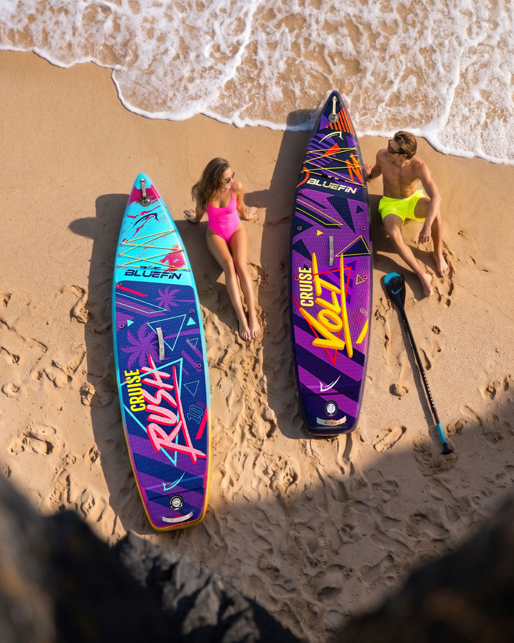 Two inflatable paddleboards on the beach, one blue and one purple, with a woman in a pink swimsuit and a man in yellow s